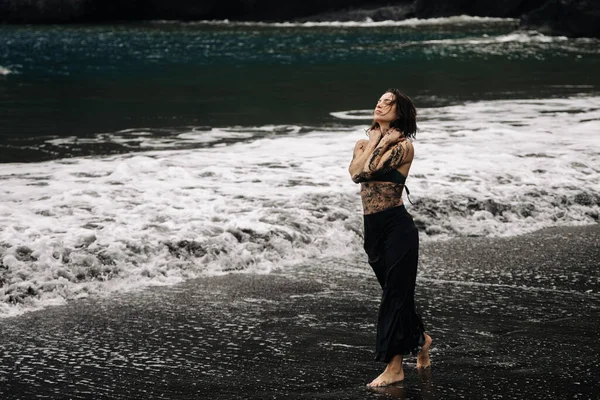 Dövmeli kadın kumlu sahilde duruyor, okyanus dalgalarını hissediyor, dalgalı siyah bir elbise giyiyor, doğal çevreyle uyum ve sükuneti somutlaştırıyor