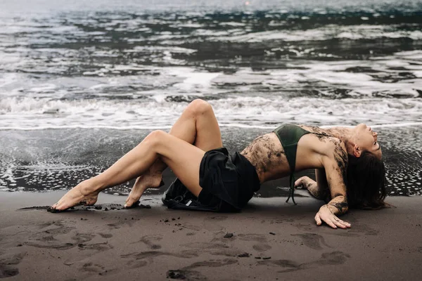 Siyah bikinili kadın figürü kumlu sahilde, çamurla süslenmiş, okyanus dalgaları plajda nazikçe tur atarken, sakin ve sanatsal bir atmosfer yaratıyor.