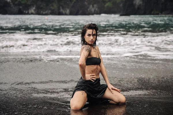 Kara kumlu sahilde diz çökmüş, okyanus dalgaları ona hafifçe çarpan, barışçıl bir atmosfer yaratan ve doğal güzelliği ve huzuru ön plana çıkaran dövmeli bir kadın modeli.