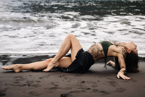 Sanatsal vücut boyalı model kumlu sahilde yatar, dalgalar ayaklarına vurur, barışçıl bir uyum ve doğanın güzelliğini yaratır.