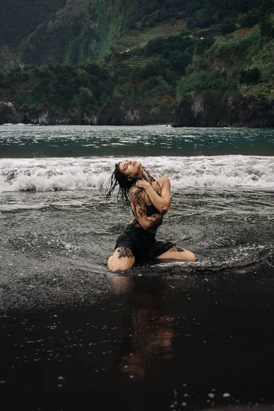 Siyah mayo giymiş kadın dansçı, okyanus dalgalarında neşeli bir şekilde hareket ediyor. Etrafına su sıçrıyor, canlı yeşil tepelere vuruyor, neşe ve özgürlüğü ifade ediyor.