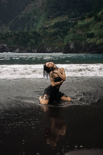 Dövmeli bir kadın, okyanus dalgalarında diz çökmüş, doğayla bağını ve özgürlüğünü somutlaştırıyor, dramatik uçurumlar ve sakin sahil manzaralarıyla çevrili, bir güç hissi uyandırıyor.