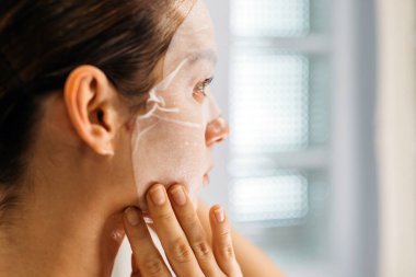 Kadın aynanın önünde yüz maskesi takıyor, cilt bakımını ve rutinini vurguluyor, hafif ışıklandırma sakin ve huzurlu bir atmosfer yaratıyor.