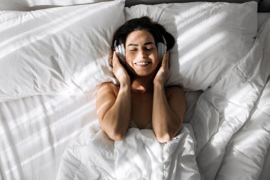 Kulaklıklı bir kadın yatakta uzanıyor, gülümsüyor ve müzikten zevk alıyor, yumuşak yastıklar ve battaniyelerle çevrili, rahat ve davetkar bir ortam yaratıyor.