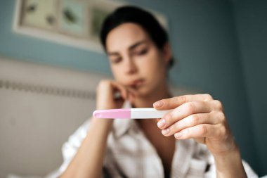 Kadın, rahat bir yatak odasında hamilelik testi yapıyor. Gelecekteki olasılıklar ve fotokopi alanındaki yaşam değişiklikleri üzerine bir anlık düşünme sergiliyor.