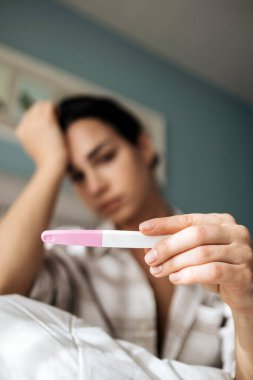 Kadın gebelik testi yapıyor. Endişeli bir bakışla, hafifçe aydınlatılmış bir odada bir yatakta oturuyor. Endişe ve potansiyel hamilelik umutlarını yansıtıyor.