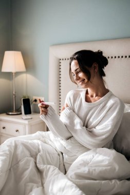 Beyaz kazaklı bir kadın, hamilelik testini neşeyle tutarak yatakta oturur, yumuşak yatak ve sıcak ışıkla rahat bir atmosfer yaratarak beklenti ve mutluluk yaratır.