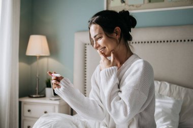 Beyaz kazaklı bir kadın yatakta oturup hamilelik testine mutlu bir şekilde bakıyor. Etrafı yumuşak yatak ve sıcak ışıklarla çevrili. Beklenti ve neşe dolu bir anı yakalıyor.