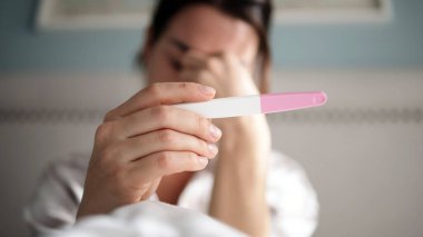 Kadın birey, yatak odasında hamilelik testi yapıyor, çeşitli duygular sergiliyor, yumuşak yatak örtüsü rahat bir düşünce ve umut atmosferi yaratıyor.