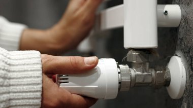 Çağdaş ortamda radyatör vanasını beyaz termostatla ayarlayan kadın, modern yaşam için ev ısıtma bakım ve enerji verimliliği uygulamalarını vurguluyor