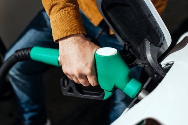 Günlük kıyafetli bir adam benzin istasyonunda yakıt ikmali yapıyor, elinde yeşil bir hortum var, yoğun bir çevrede araçlara yakıt ikmali yapıldığını gösteriyor.