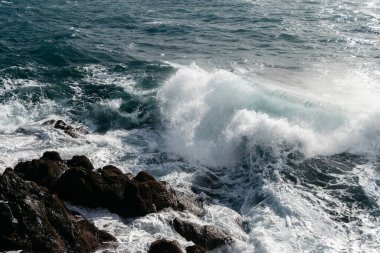 Dalgalar kıyı şeridi boyunca kayalara çarpar, köpük ve su sıçratır, güneş ışığı suda parıldar, doğanın güzellik ve enerjisinin özünü yakalar.