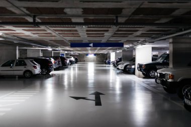 Yeraltı otoparkında birçok park edilmiş araç yer alıyor, yukarıdaki ışıklarla aydınlatılıyor, modern tasarım ve kentsel kolaylık vurgulanıyor.