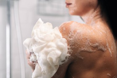 Koyu renk saçlı kadın, duşta omzuna köpüklü sabun köpüğü sarılıyor. Yumuşak ışıklandırma ve sakin bir ortam ile ferahlatıcı bir banyo deneyimini vurguluyor.