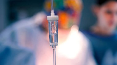 Temiz sıvıyla dolu damar içi damlalar tıbbi bir ortamda belirgin bir şekilde sergilenirken, arka planda sağlık uzmanları bulanıklaşarak hasta bakımının önemini vurguluyor.