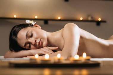 Asyalı kadın, bir masaj masasında huzur içinde uzanıyor. Yumuşak mum ışığı ve nazik dekorla sarmalanmış, rahatlama ve sükuneti teşvik eden huzurlu bir atmosferde.