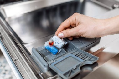 Bir el bulaşık makinesine deterjan tableti yerleştiriyor, modern bir mutfak ortamında temizlik sürecini vurguluyor, kolaylık ve verimliliği vurguluyor.