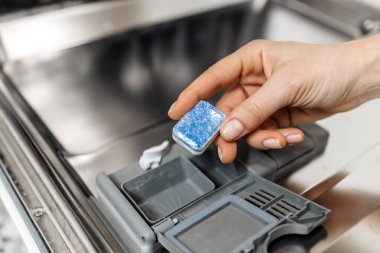 Close-up of hand placing blue dishwasher tablet into open compartment of modern dishwasher, highlighting functionality and ease of use in kitchen cleaning tasks
