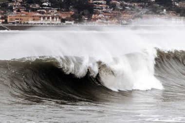 Görkemli okyanus dalgası kumlu sahile çarpıyor, uzak bir sahil kasabası doğanın dinamik enerjisini yakalayarak sakin bir zemin oluşturuyor.