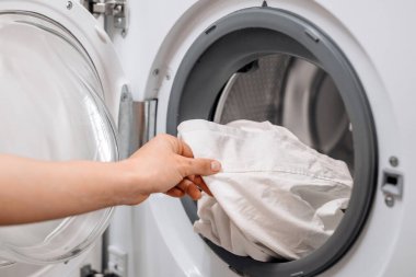 Bireyin biri çamaşır makinesinden temiz beyaz bir gömlek çıkarıyor. Çamaşır makinesinin parlak ve düzenli bir çamaşır odasında çamaşırların yıkanma sürecini vurguluyor.