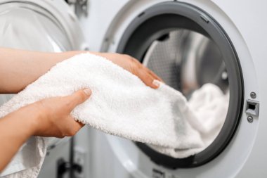 Kurutucudan yumuşak beyaz havluyu çıkaran kişi, modern aletler ve temiz bir atmosferle aydınlık, düzenli bir çamaşır odasında çamaşırları temizliyor.