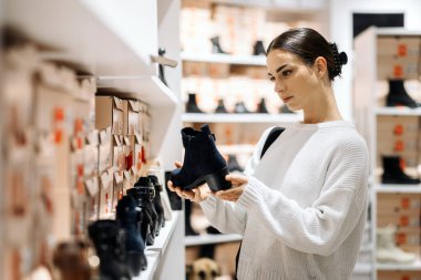 Yumuşak bir süveter giyen bayan alışverişçi, çeşitli ayakkabı seçenekleriyle dolu raflarla çağdaş bir ayakkabı mağazasında modaya uygun siyah ayak bileği botlarını denetliyor.