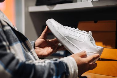 Mağazada modaya uygun beyaz bir spor ayakkabı tutan kişi, arka planda canlı ayakkabı kutuları, çağdaş moda ve perakende atmosferi vurguluyor.