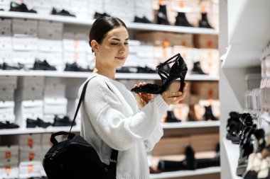 Çağdaş bir mağazada siyah yüksek topuklu ayakkabı satan kadın, arka planda çeşitli ayakkabı rafları, moda tercihleri ve perakende ambiyansı vurguluyor.