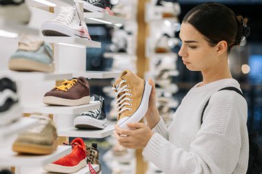 Kadın tüketici, modern bir mağazada moda ayakkabılarını yakından inceliyor, çeşitli ayakkabılar sergileniyor, moda tercihlerini ve tüketici katılımını vurguluyor