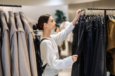 Koyu renk saçlı bir alışverişçi şık bir perakende piyasasında elbise raflarını keşfediyor, moda eğilimlerini ve kişisel tercihlerini ön plana çıkarıyor.