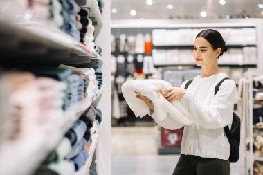 Şık bir süveter giyen kadın alışverişçi, şık bir perakende alanında havluları seçiyor, renkli tekstil ürünleri raflarda sergileniyor ve canlı bir atmosfer yaratıyor.