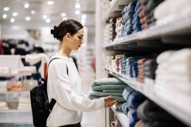 Beyaz kazaklı bir kadın alışverişçi, çağdaş bir mağazanın rafındaki havlulara göz atıyor, perakende ortamı ve ürün seçimini vurguluyor.