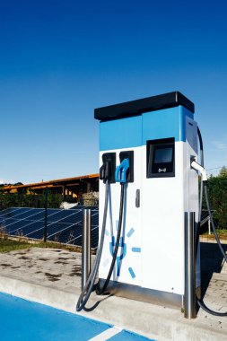 Elektrikli araç şarj istasyonu, yenilenebilir enerji ve yeniliğin vurgulandığı açık mavi gökyüzü ve güneş panellerinden oluşan bir zemin üzerine kurulmuş iki şarj bağlantısına sahip.