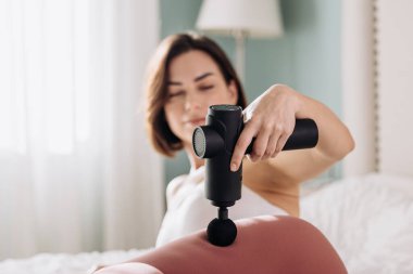 Kadın birey yatakta otururken sırtına titreşimli masaj tabancası uyguluyor. Yumuşak ışıklandırmayla sakin bir ortam yaratıyor ve rahatlama ve sağlığa odaklanıyor.