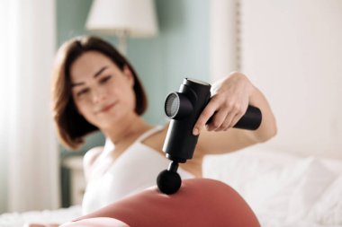 Kadın bir yatakta dinlenirken kalçasına titreşimli masaj tabancası uyguluyor. Kas gevşeme ve iyileşme tekniklerinin rahatlatıcı etkilerini vurguluyor.