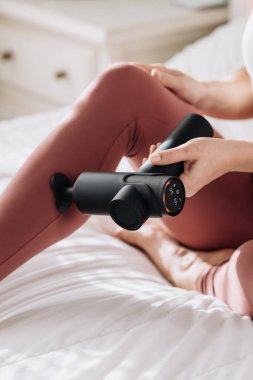 Yatakta otururken kalçasında titreşimli masaj tabancası kullanan bir kadın, yumuşak dokuları olan huzurlu bir evde rahatlık ve rahatlama vurguluyor.