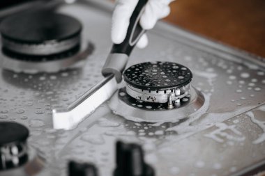 Eldiven içinde bir gaz sobasını fırçayla ovmak, parlak yüzeyleri ve su damlacıklarını ortaya çıkarmak, temizliği ve mutfak bakımını vurgulamak,