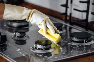 Eldivenli el paslanmaz bir çelik gaz sobasını süngerle ovuyor. Mutfaktaki temizlik ve hijyeni vurguluyor. Ev işlerini ve bakım masraflarını vurguluyor.