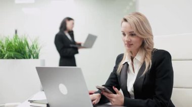 Profesyonel bir toplantı sırasında arka planda dizüstü bilgisayarları ve cep telefonlarıyla modern bir ofis masasında çalışan iki iş kadını.