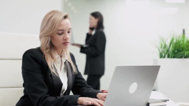 Profesyonel bir toplantı sırasında arka planda dizüstü bilgisayarları ve cep telefonlarıyla modern bir ofis masasında çalışan iki iş kadını.