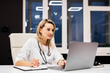 Steteskoplu kadın doktor, modern bir ofis masasında bilgisayarına odaklanmış. Yanında not defteri ve kalem var. Arka planda büyük pencereler var.