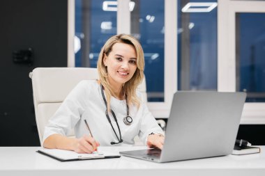 Kadın doktor bir masada oturuyor, notlar yazıyor ve modern bir ofiste geniş pencereli ve çağdaş dekorlu bir dizüstü bilgisayar kullanıyor.