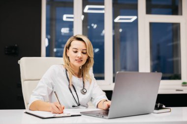 Kadın doktor, akşamları büyük pencereli ve çağdaş ışıklandırmalı modern bir ofis masasında dizüstü bilgisayar kullanırken notlar yazıyor.