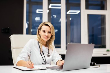 Steteskoplu kadın doktor bir masada oturuyor, dizüstü bilgisayar kullanıyor ve modern bir ofiste büyük pencereleri ve parlak ışıklandırması olan notlar yazıyor.