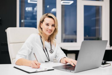 Steteskoplu kadın doktor bir masada oturuyor, dizüstü bilgisayar kullanıyor ve modern bir ofiste büyük pencereleri ve şık dekorasyonu olan notlar yazıyor.