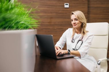 Beyaz önlüklü kadın doktor, profesyonel bir ayarlama sırasında arka planda yeşil bitkiler olan şık bir ofis masasında dizüstü bilgisayar kullanıyor.