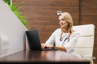 Beyaz önlüklü kadın doktor, çağdaş bir sağlık ofisinde yeşil bitkiler ve ahşap bir duvarla şık bir masada dizüstü bilgisayar kullanıyor.