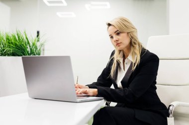 Siyah takım elbiseli iş kadını, arka planda yeşil bitkiler ve çağdaş ışıklandırmayla süslü bir ofis masasında dizüstü bilgisayarına odaklanıyor.