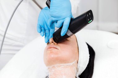 Güzellik kliniğinde tedavi gören bir kadın. Teknisyen, steril bir ortamda tedavi yatağında yatarken yüzüne cihaz uyguluyor.