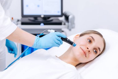 Kadın kozmetik uzmanı, tedavi yatağında yatan bir hastaya hidrakiyal prosedür uyguluyor. Tıbbi cihaz ve monitör, parlak bir klinik ortamda görülüyor.